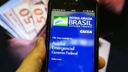 Pessoa segura dinheiro em espécie enquanto consulta aplicativo de benefício social da Caixa Econômica Federal no celular