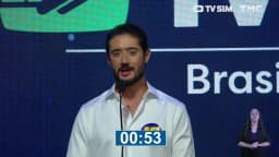 Gabriel Azevedo, candidato do MDB ao governo de Minas Gerais, discursa em pé no púlpito durante o debate, usando camisa branca com o número 15, com o telão da TV Sim e da TMC ao fundo e intérprete de Libras ao canto