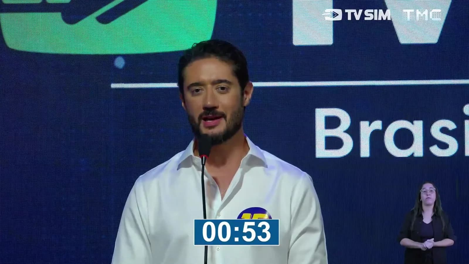 Gabriel Azevedo, candidato do MDB ao governo de Minas Gerais, discursa em pé no púlpito durante o debate, usando camisa branca com o número 15, com o telão da TV Sim e da TMC ao fundo e intérprete de Libras ao canto