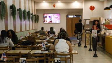 Clientes às mesas de um restaurante em funcionamento, com balcão de buffet e atendentes ao fundo