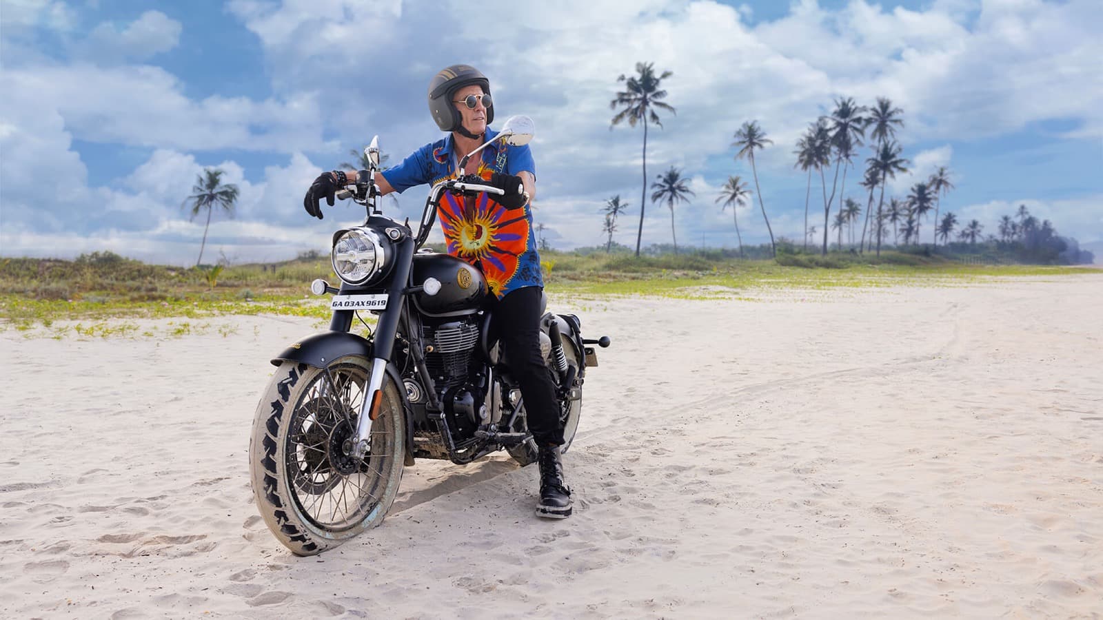 Motociclista sentado sobre uma Royal Enfield Goan Classic 350 preta em uma praia de Goa, na Índia, com coqueiros ao fundo. Imagem de divulgação da fabricante.
