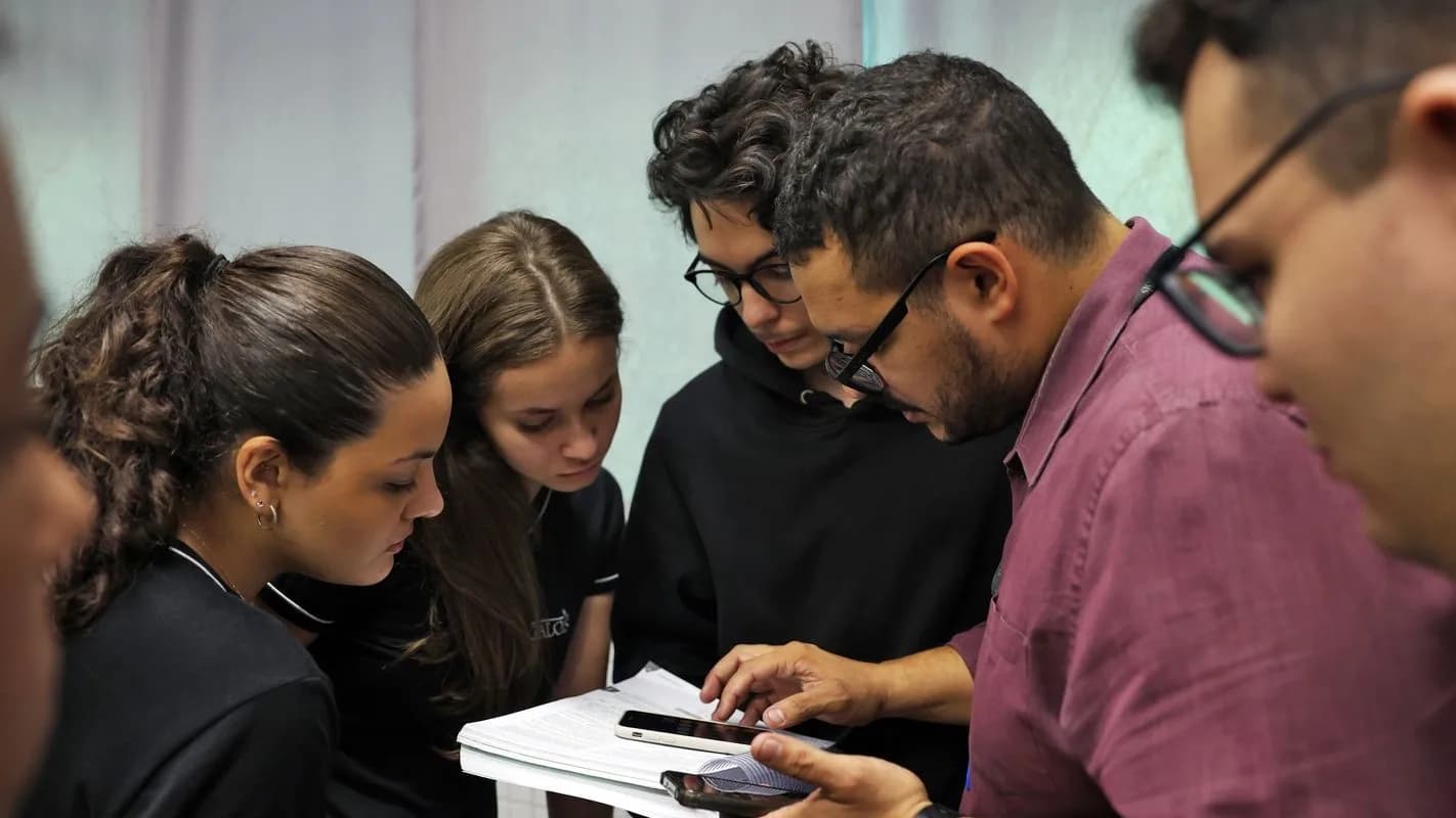 Professor auxilia grupo de estudantes que observam documento e celular durante atividade em sala