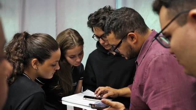 Professor auxilia grupo de estudantes que observam documento e celular durante atividade em sala