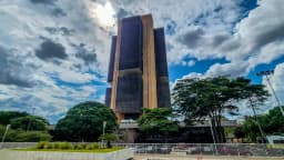 Fachada da sede do Banco Central do Brasil em Brasília, prédio revestido em vidro escuro e ripas douradas, sob céu com nuvens