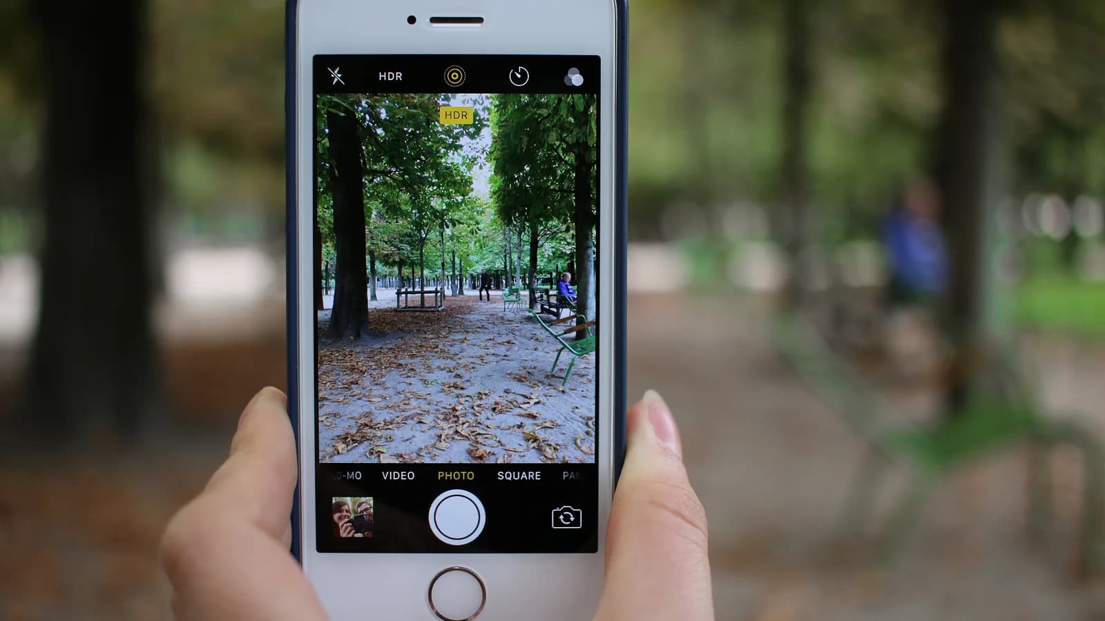 Pessoa segura um iPhone com as duas mãos para fotografar um parque (foto ilustrativa de arquivo, modelo anterior ao 18 Pro)