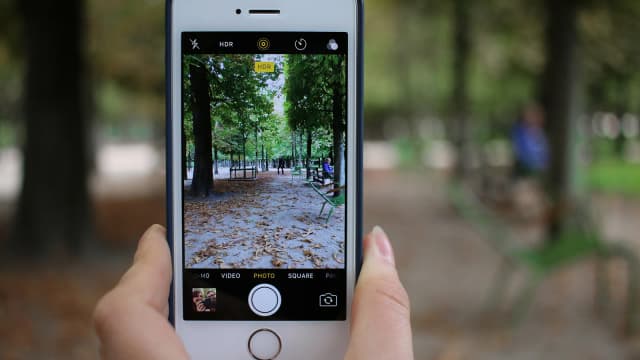 Pessoa segura um iPhone com as duas mãos para fotografar um parque (foto ilustrativa de arquivo, modelo anterior ao 18 Pro)