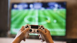 Mãos seguram controle de videogame enquanto jogam partida de futebol na tela