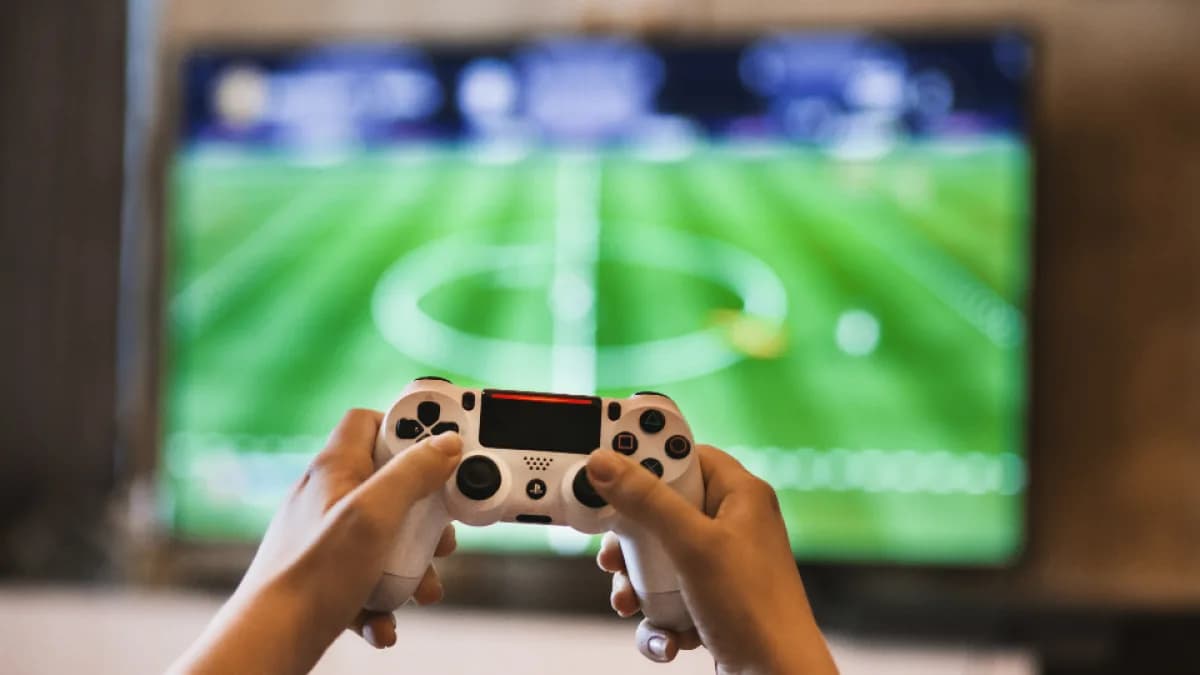 Mãos seguram controle de videogame enquanto jogam partida de futebol na tela