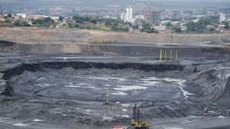 Vista aérea de mina a céu aberto no Brasil, com maquinário e caminhões de mineração ao fundo de área urbana