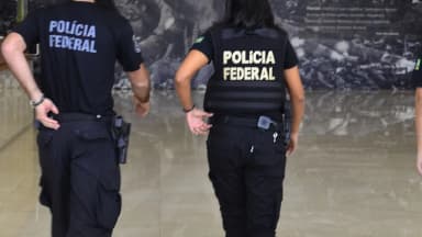 Agentes da Polícia Federal, vistos de costas, com o colete e a camisa identificados com o nome da corporação