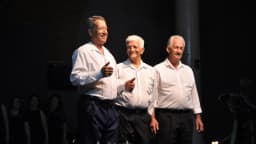 Três idosos sorridentes em pé, de camisa branca, em evento social para a terceira idade