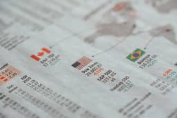 Página de jornal econômico mostra cotações de bolsas de valores com as bandeiras do Brasil, Estados Unidos e Canadá, ilustrando o impacto das tarifas dos EUA sobre o mercado brasileiro