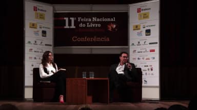 Augusto Cury fala ao microfone durante conferência em feira de livro, sentado ao lado de entrevistadora