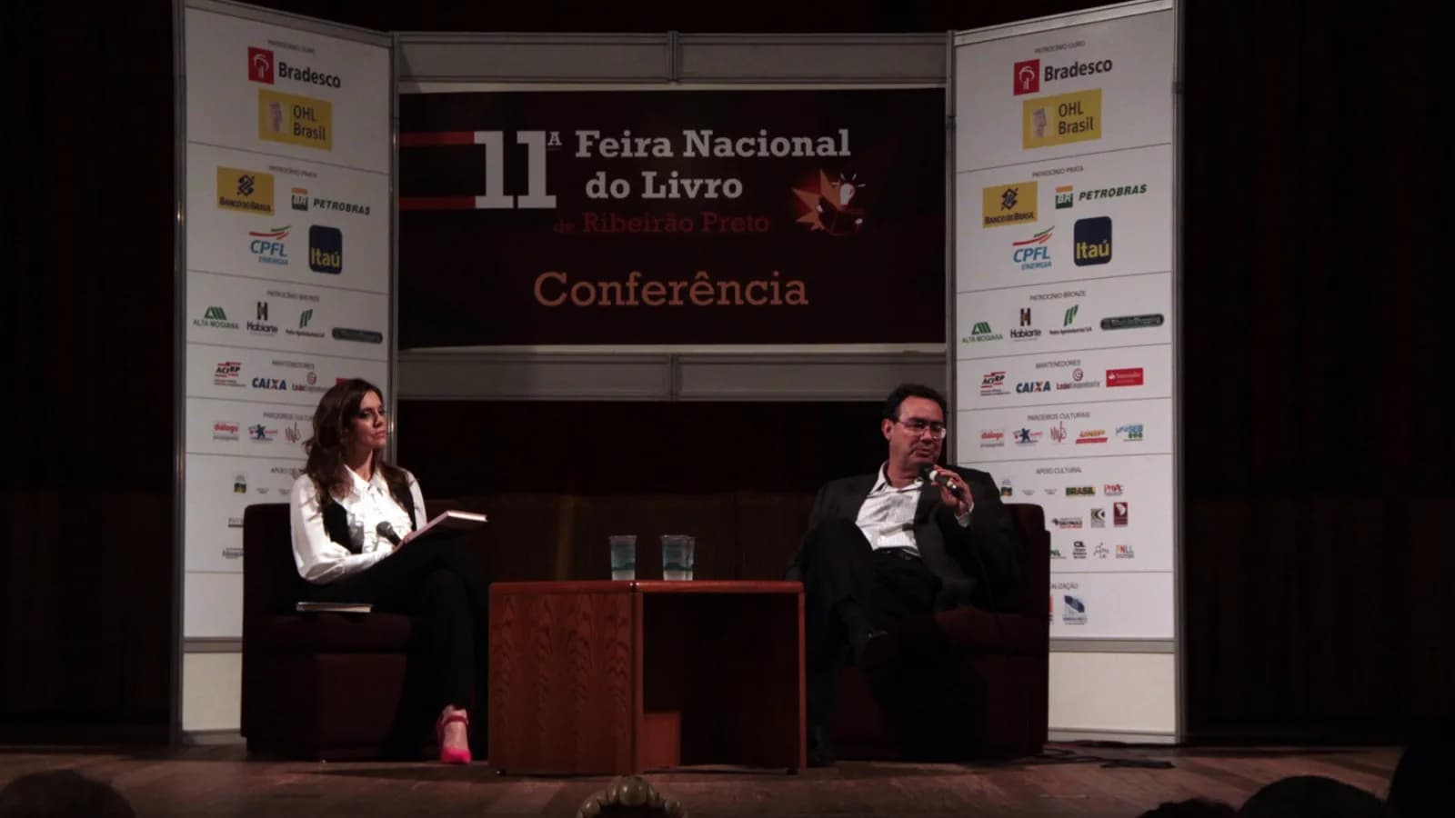 Augusto Cury fala ao microfone durante conferência em feira de livro, sentado ao lado de entrevistadora