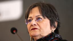 Maria da Penha, de óculos e cachecol estampado, em evento público falando ao microfone