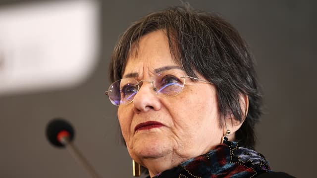 Maria da Penha, de óculos e cachecol estampado, em evento público falando ao microfone