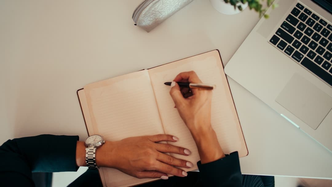Pessoa escreve à mão em um caderno ao lado de um notebook aberto (foto ilustrativa)