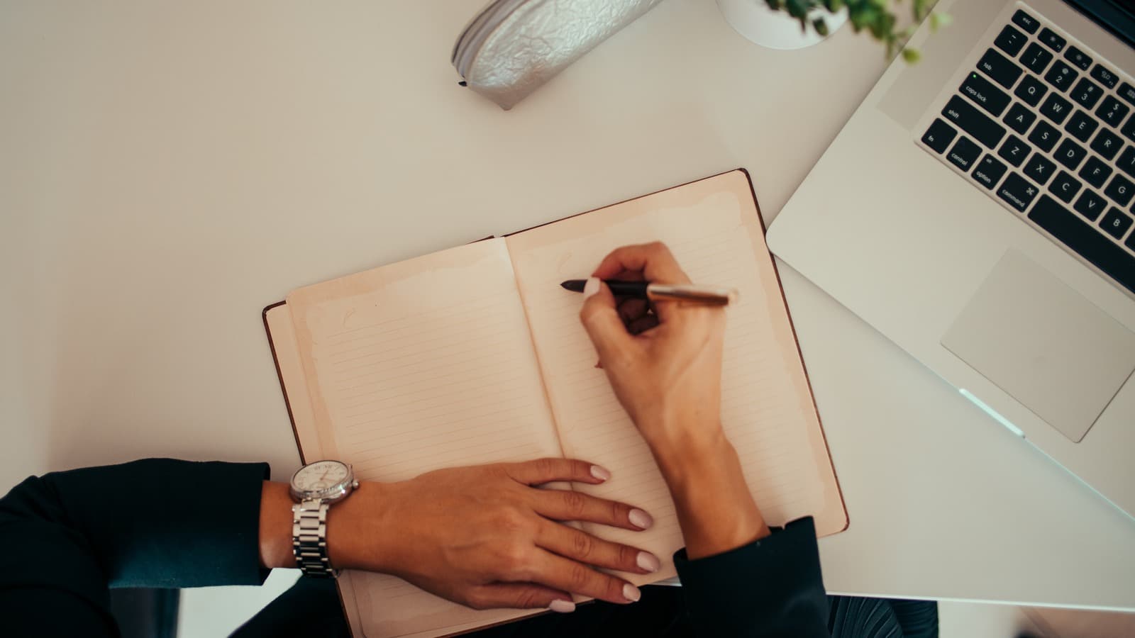 Pessoa escreve à mão em um caderno ao lado de um notebook aberto (foto ilustrativa)