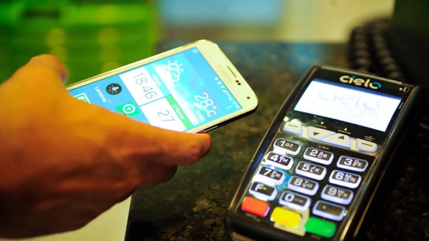 Mão segura celular próximo a máquina de cartão, em pagamento por aproximação