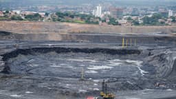 Vista de ampla mina a céu aberto com bancadas escavadas, sonda de perfuração e máquinas ao fundo de uma área urbana; imagem de arquivo