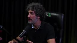 Carlos Eduardo Grimaldi gesticula com a mão enquanto fala, olhando em direção à entrevistadora, em estúdio de podcast com cortina escura ao fundo