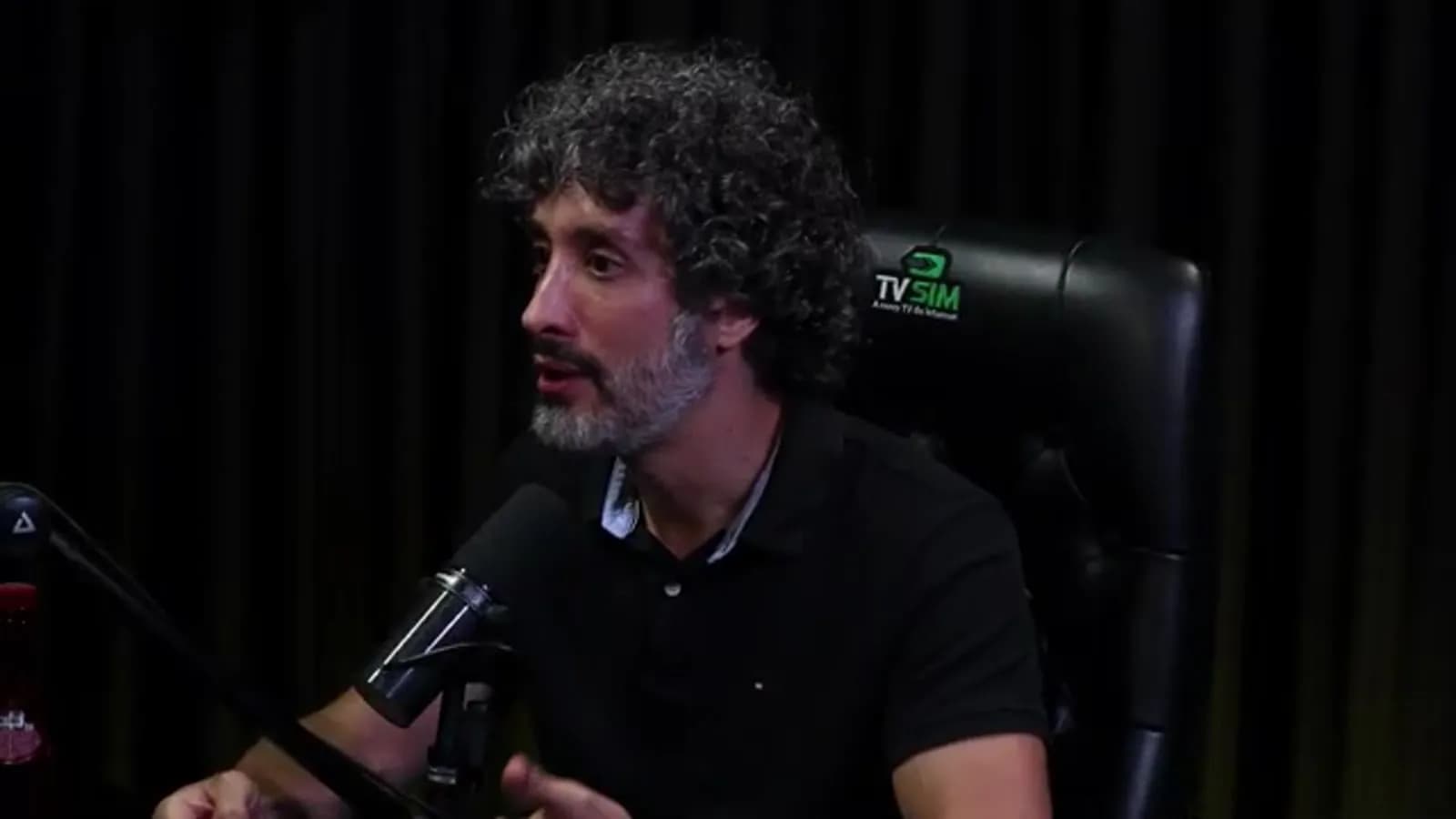 Carlos Eduardo Grimaldi gesticula com a mão enquanto fala, olhando em direção à entrevistadora, em estúdio de podcast com cortina escura ao fundo