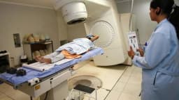 Paciente em sessão de radioterapia oncológica, com profissional de saúde operando o equipamento, em foto de arquivo