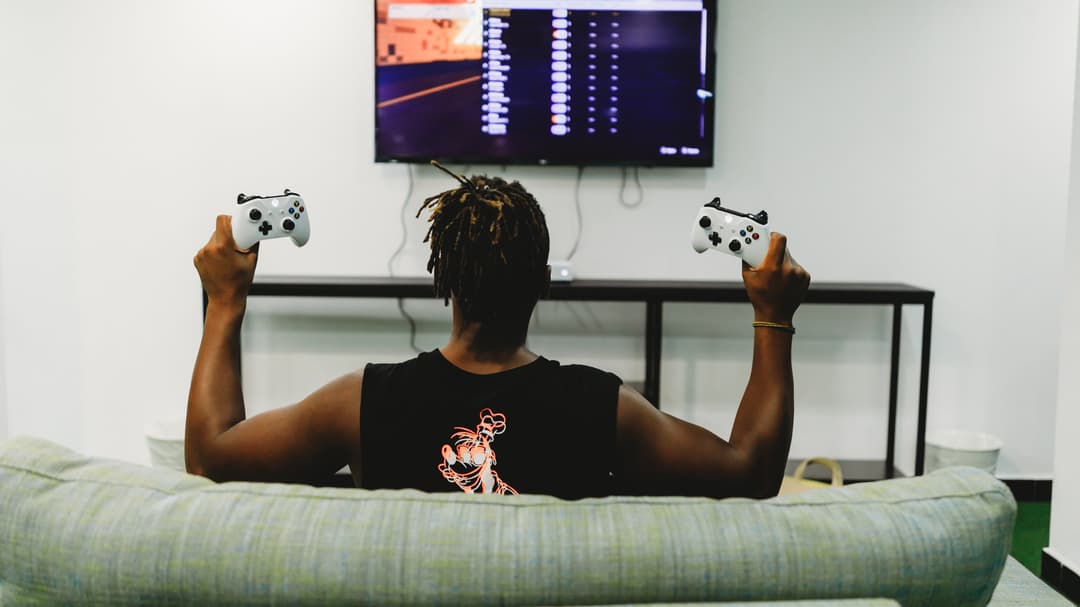 Pessoa de costas joga videogame no sofá segurando dois controles de Xbox (foto ilustrativa)
