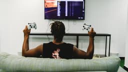 Pessoa de costas joga videogame no sofá segurando dois controles de Xbox (foto ilustrativa)