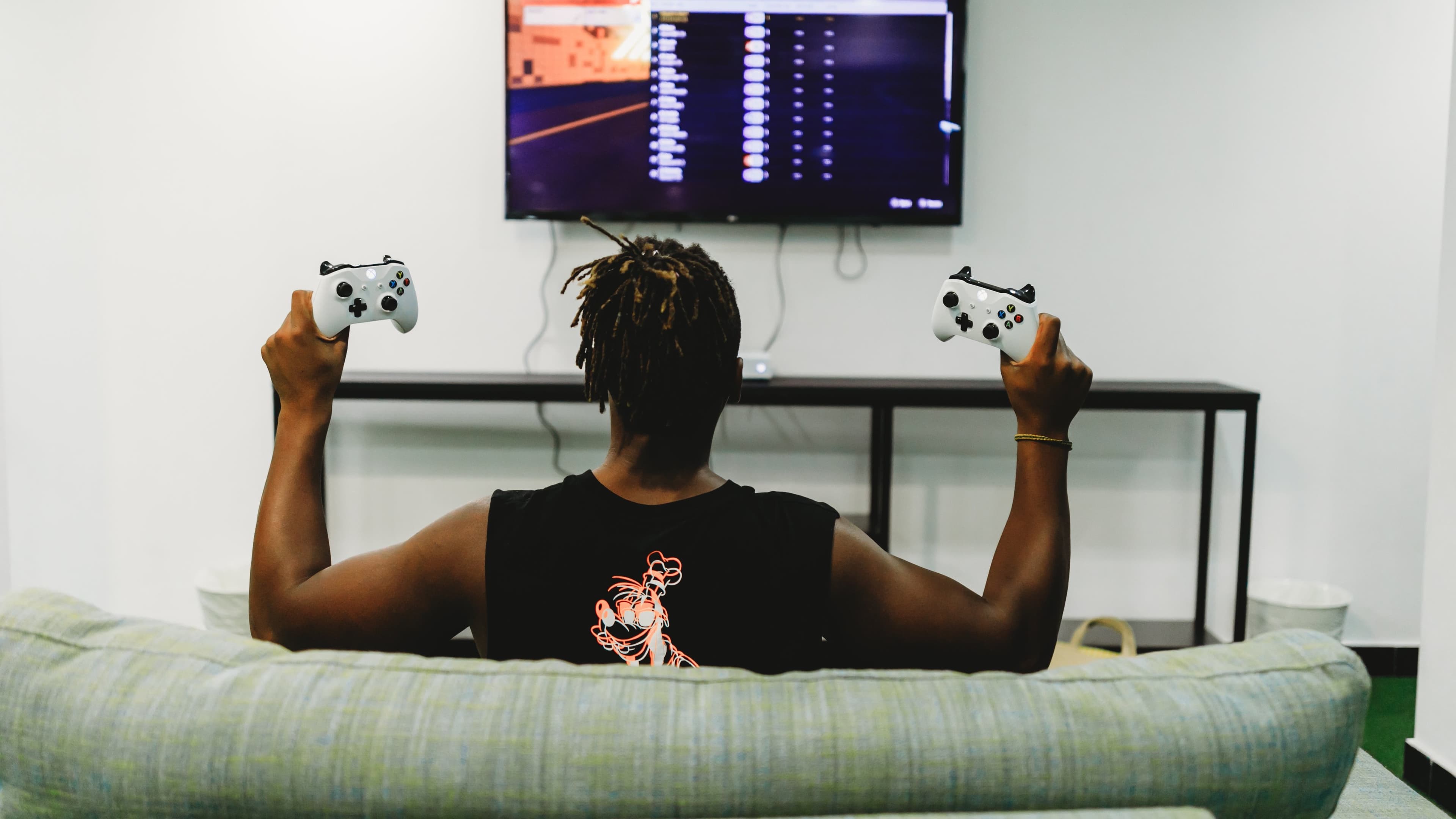 Pessoa de costas joga videogame no sofá segurando dois controles de Xbox (foto ilustrativa)