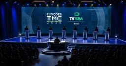 Palco do debate com oito púlpitos iluminados, telão com as marcas Eleições TMC 2026 e TV Sim Brasil e o mediador de costas ao centro, diante da plateia.