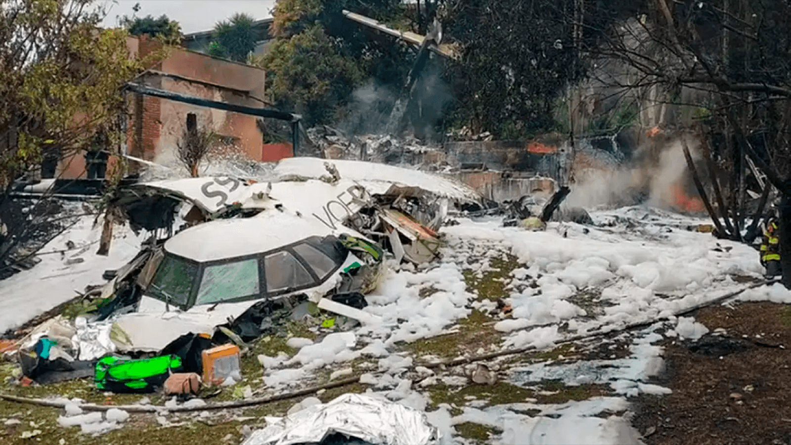 Destroços do avião da Voepass após a queda em Vinhedo (SP), com bombeiro ao fundo cercado por espuma de combate a incêndio