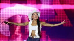 Ana Castela durante apresentação em show, com chapéu branco e microfone