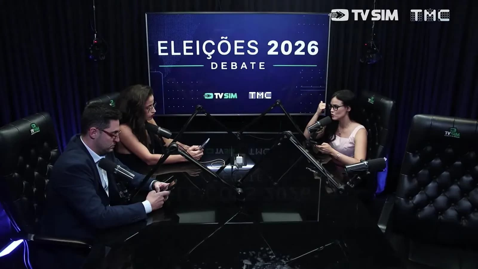 Apresentadores em mesa redonda de estúdio, com telão ao fundo mostrando 'Eleições 2026 Debate', logos da TV Sim e da TMC