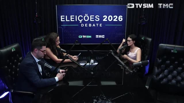 Apresentadores em mesa redonda de estúdio, com telão ao fundo mostrando 'Eleições 2026 Debate', logos da TV Sim e da TMC