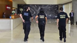 Três agentes da Polícia Federal caminham em corredor durante ação, uniformizados com a inscrição Polícia Federal