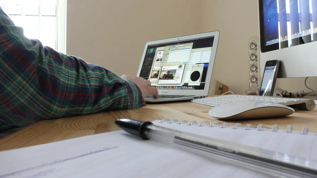 Pessoa trabalhando em um laptop sobre mesa de madeira, com caderno e caneta ao lado