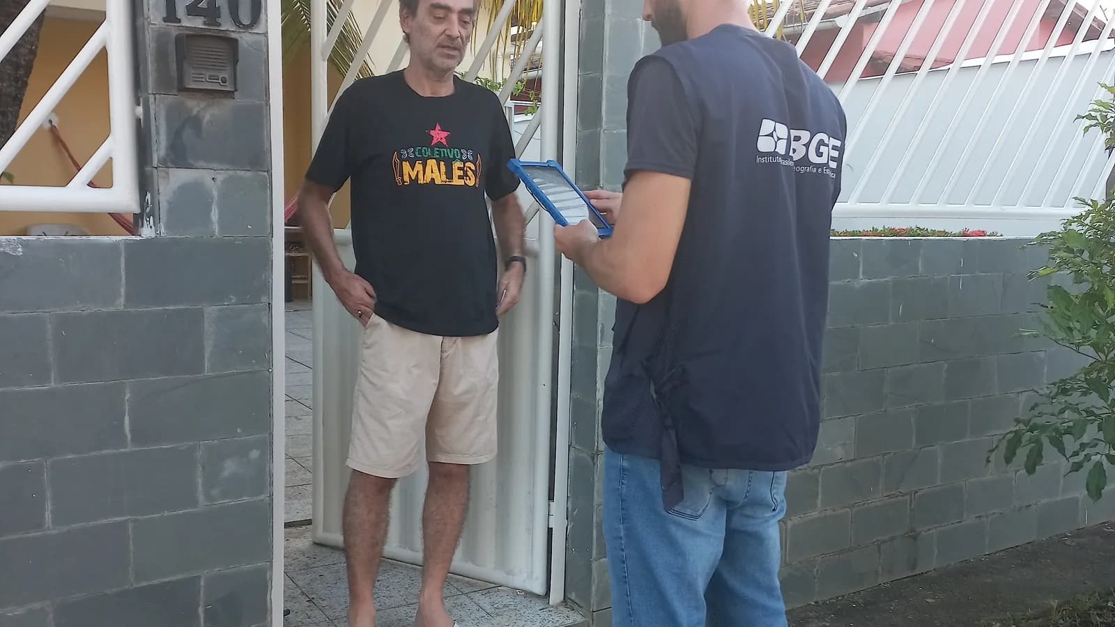 Recenseador do IBGE, identificado por camisa com a logo do instituto, entrevista morador em visita domiciliar