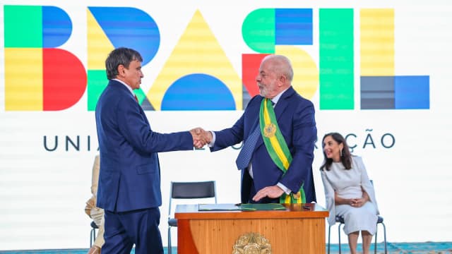 Lula aperta a mão do ministro Wellington Dias em cerimônia no Palácio do Planalto, com painel escrito Brasil ao fundo