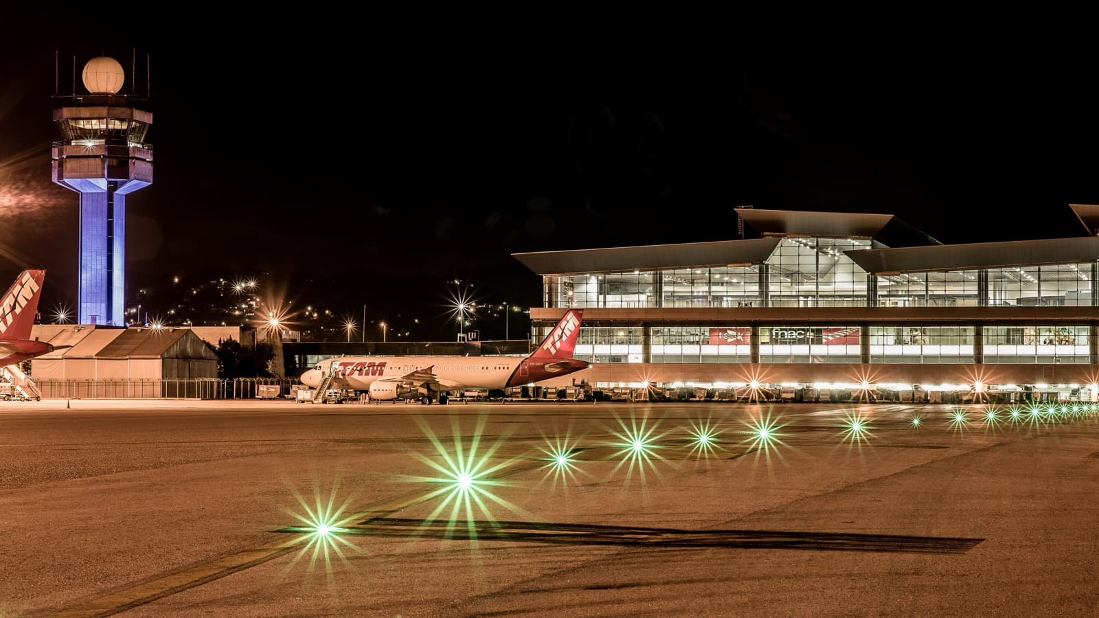 Pátio do Aeroporto Internacional de São Paulo, em Guarulhos, à noite, com a torre de controle iluminada, o terminal de passageiros ao fundo e luzes verdes de balizamento na pista. Imagem de arquivo.