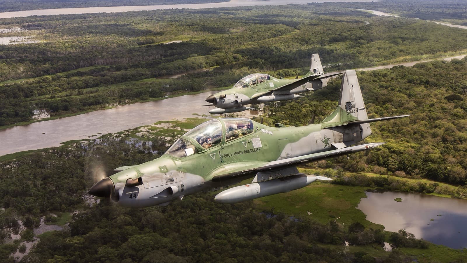 Dois caças A-29 Super Tucano da Força Aérea Brasileira voando lado a lado sobre área de floresta e rio, em imagem de arquivo