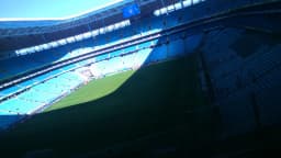 Arquibancada lotada da Arena do Grêmio durante partida, com torcida tricolor e gramado ao fundo