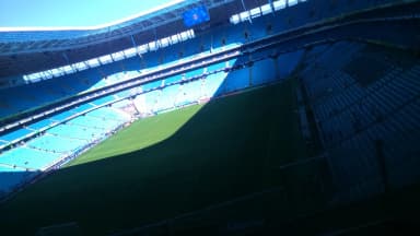 Arquibancada lotada da Arena do Grêmio durante partida, com torcida tricolor e gramado ao fundo