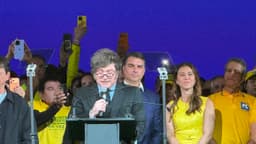 Javier Milei discursa ao microfone durante convenção do PL em São Paulo, com Flávio Bolsonaro ao fundo