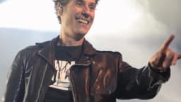 Dinho Ouro Preto sorri no palco, usando jaqueta de couro preta, apontando para o público durante show