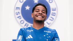 Atacante Wesley sorri com a camisa do Cruzeiro em frente ao escudo do clube, durante apresentação oficial