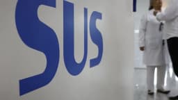 Logo do SUS em parede de unidade de saúde, com profissionais de jaleco branco ao fundo
