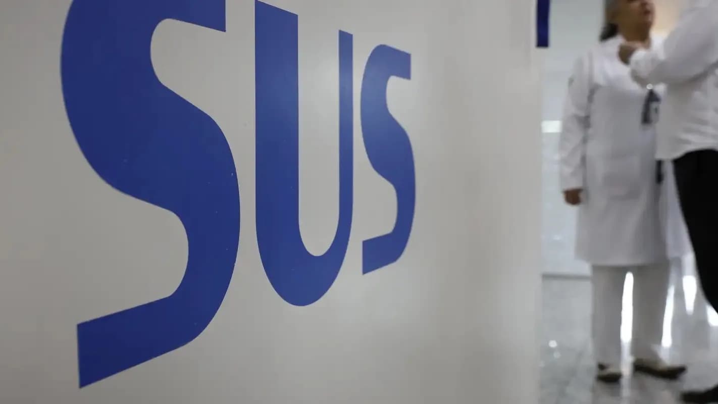 Logo do SUS em parede de unidade de saúde, com profissionais de jaleco branco ao fundo