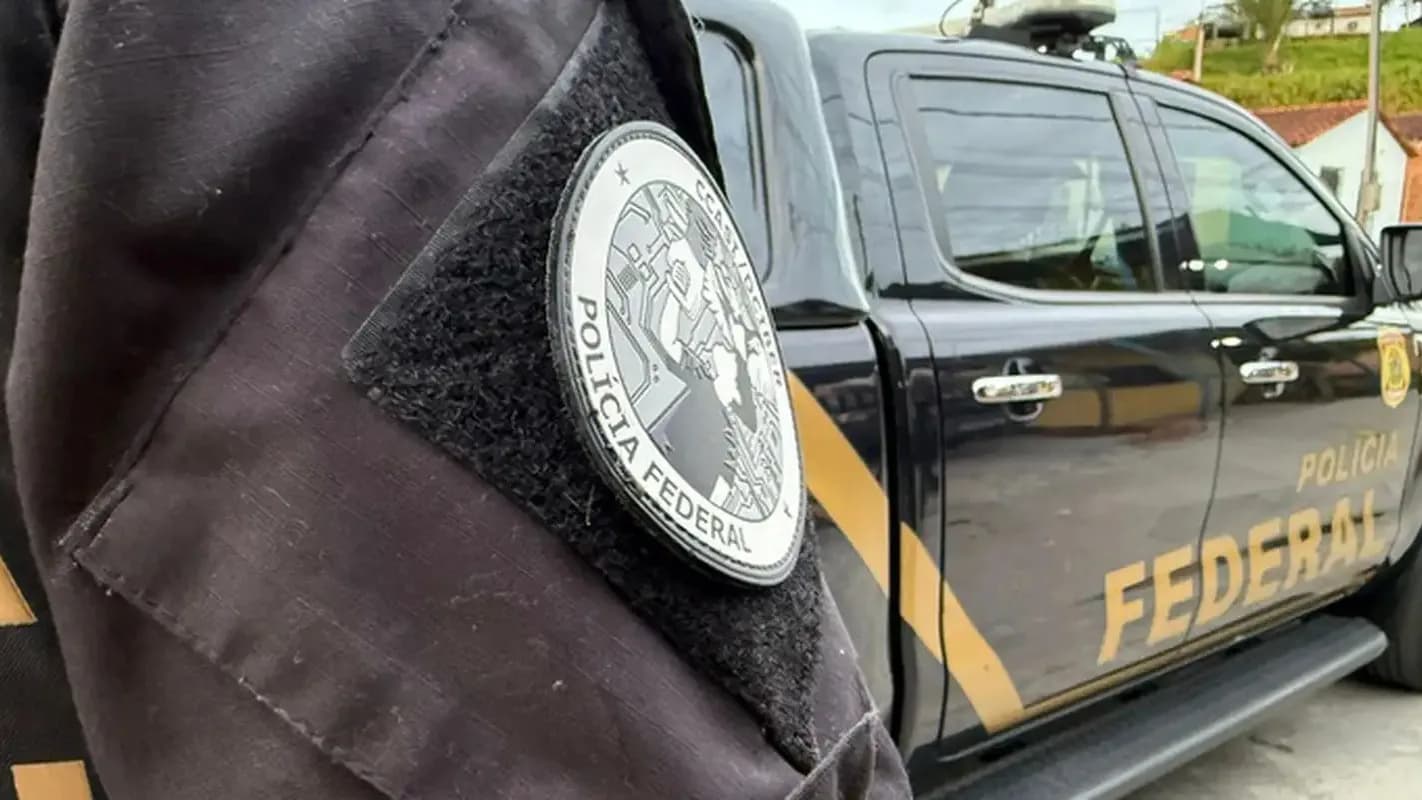 Distintivo da Polícia Federal em uniforme de agente ao lado de viatura, em operação contra o Comando Vermelho