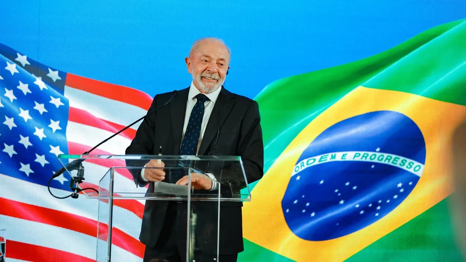 O presidente Lula sorri em coletiva de imprensa em Washington, com bandeiras dos Estados Unidos e do Brasil ao fundo